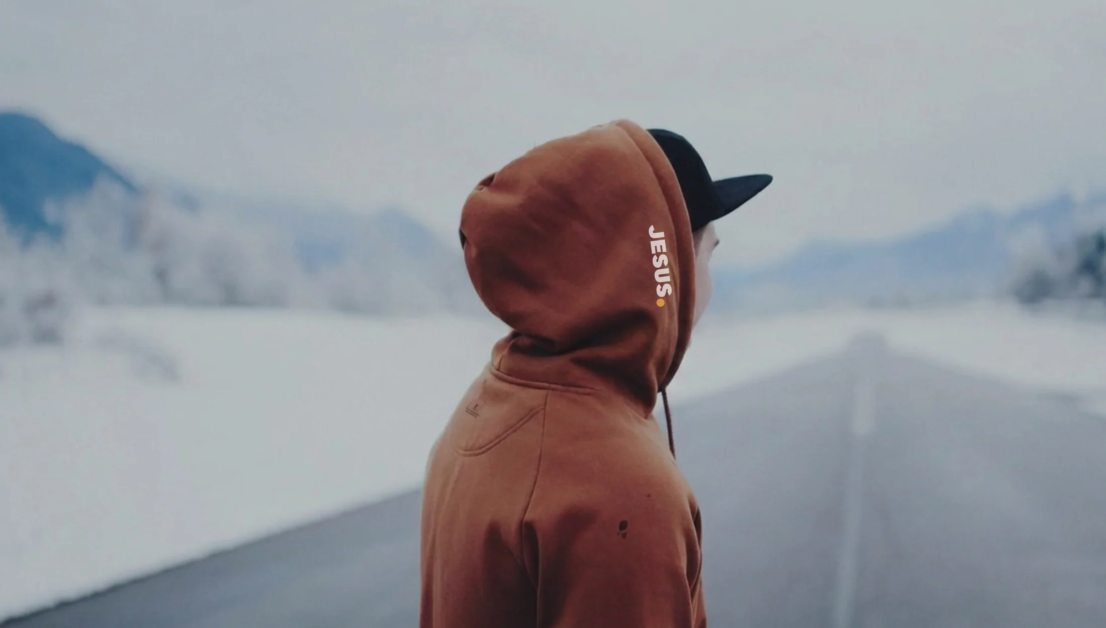 Person wearing a rust Jesus Period Christian hoodie beside a snowy mountain lake